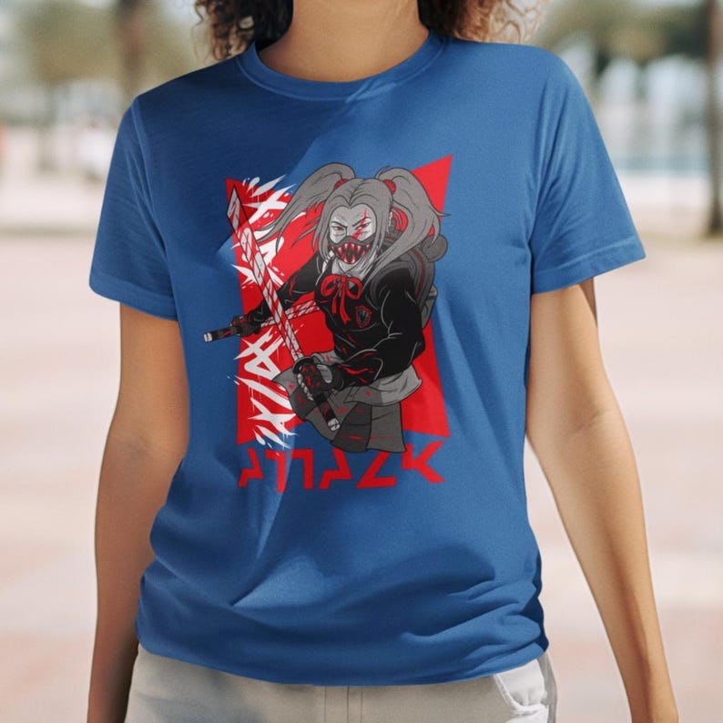 May include: A royal blue t-shirt with a graphic of a girl with pigtails wearing a black hooded jacket and holding a sword. The girl has a red and white mask on her face. The graphic is set against a red background with white jagged lines and the word 'ATTACK' written in white letters below the graphic.