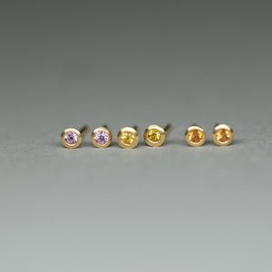K&ouml;nnte beinhalten: F&uuml;nf Paar goldene Ohrstecker mit kleinen Edelsteinen. Die Edelsteine sind rosa, hellrosa, gelb, orange und hellorange.