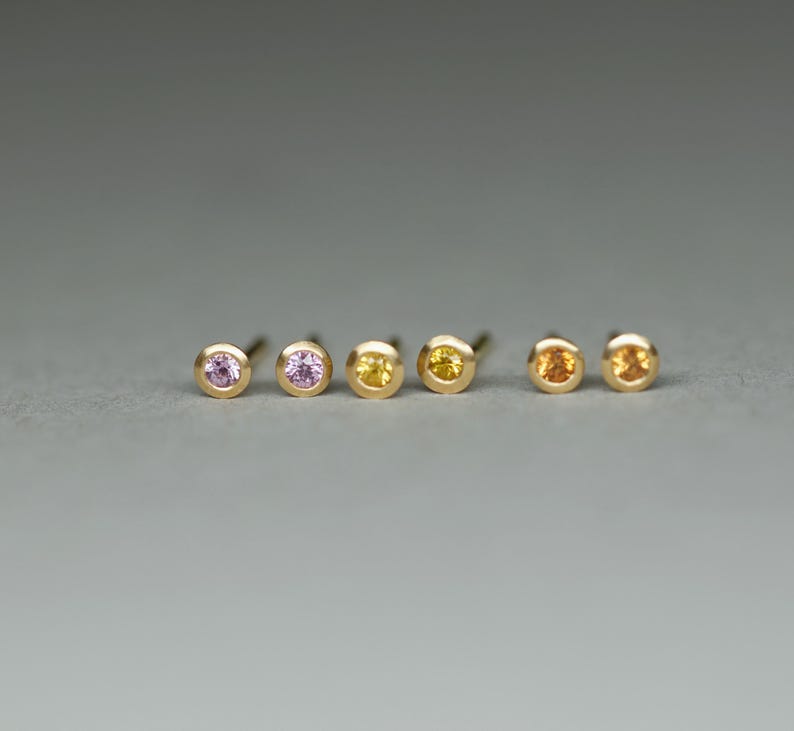 K&ouml;nnte beinhalten: F&uuml;nf Paar goldene Ohrstecker mit kleinen Edelsteinen. Die Edelsteine sind rosa, hellrosa, gelb, orange und hellorange.