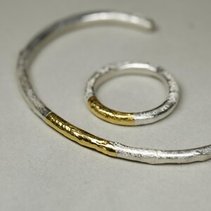 May include: A set of two silver and gold rings. The larger ring is a bangle with a gold band on one side. The smaller ring is a simple band with a gold band on the outside.