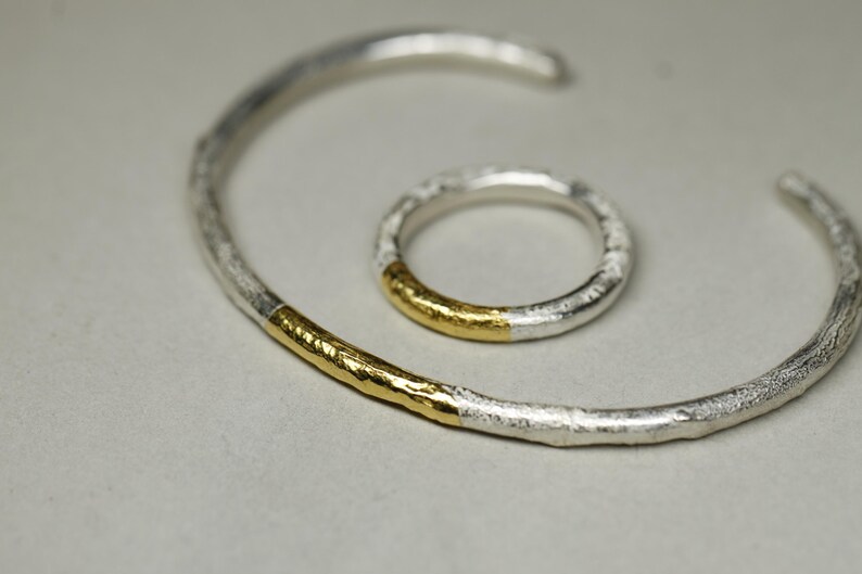 May include: A set of two silver and gold rings. The larger ring is a bangle with a gold band on one side. The smaller ring is a simple band with a gold band on the outside.