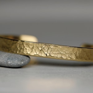 Könnte beinhalten: Ein goldfarbenes, gehämmertes Armkettchen. Das Armband besteht aus dickem, strukturiertem Metall und hat ein schlichtes, elegantes Design.