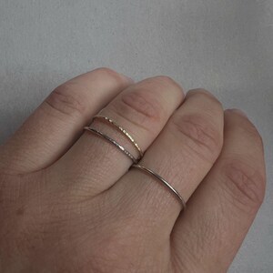 May include: Two thin gold rings stacked on a finger. The top ring has a hammered texture, while the bottom ring is smooth.