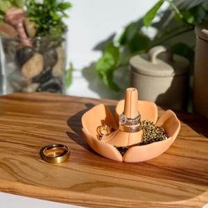 May include: A peach-colored, flower-shaped ring holder with a central post and petal-like compartments. The holder displays various rings and a gold chain. A gold ring rests on a wooden tray, alongside a small glass with plants.