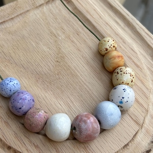 May include: A necklace featuring a string of colorful, speckled beads in shades of blue, purple, pink, white, and orange. The beads are round and vary in size, strung on a green cord. The necklace is displayed on a wooden surface.