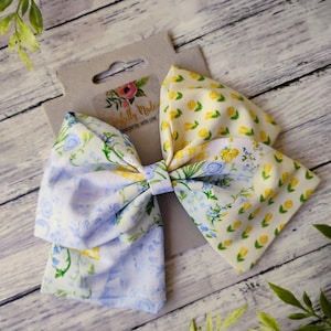 May include: A large fabric bow with a floral pattern in shades of blue, yellow, and green. The bow is attached to a cardboard tag with the text "Joyfully Made, handcrafted with love". The bow is on a wooden surface.