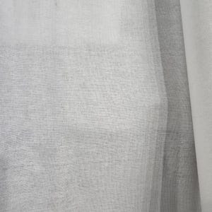 May include: Close-up of a sheer, light gray fabric with a woven texture. The fabric is slightly translucent, allowing some light to pass through. The material appears to be lightweight and possibly linen or a similar textile.