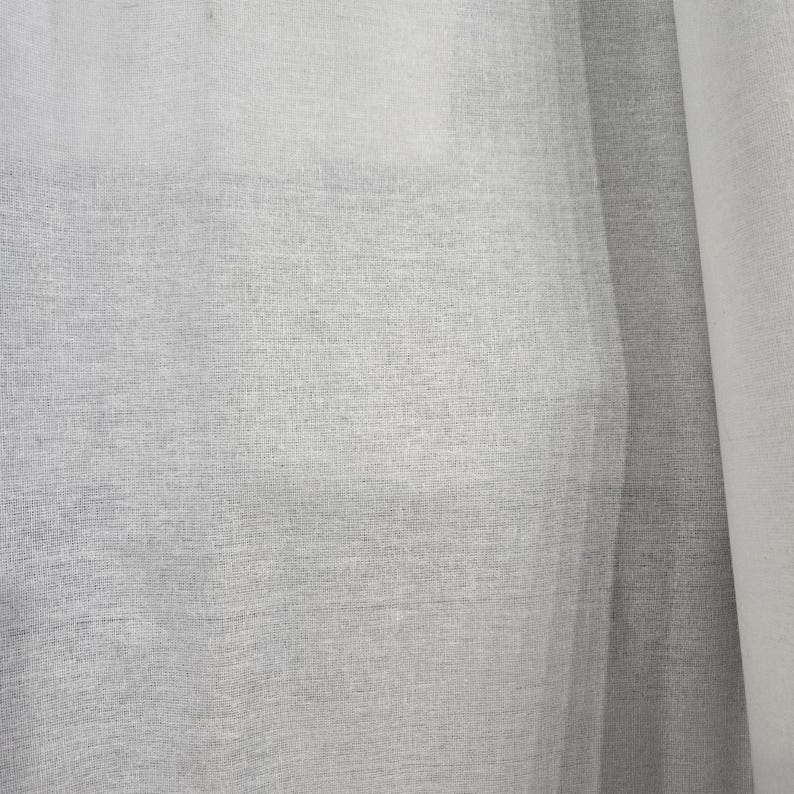 May include: Close-up of a sheer, light gray fabric with a woven texture. The fabric is slightly translucent, allowing some light to pass through. The material appears to be lightweight and possibly linen or a similar textile.