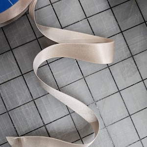 Puede incluir: Cinta de raso beige desenrollándose sobre una superficie de cuadrícula gris. La cinta es lisa y tiene un ligero brillo, con un diseño curvo y fluido. Adecuada para manualidades, costura o envolver regalos.