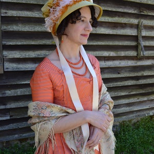 以下が含まれることがあります： オレンジと白のストライプのヴィンテージスタイルのドレスを着た女性。白いショールとオレンジと白の羽根のついた黄色のボンネットを被っている。彼女は木製の壁の前に立っている。