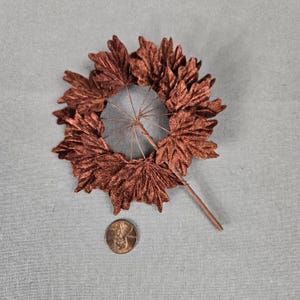 May include: A cluster of metallic copper-colored artificial maple leaves attached to thin wire stems, arranged in a circular fan shape. A single United States penny is positioned below the leaves to provide a clear scale for the size of the object.