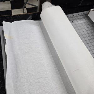 May include: A roll of white fabric, partially unrolled, lies on a cutting mat. The fabric appears to be a lightweight material, possibly cotton or linen. The scene suggests a sewing or crafting project.