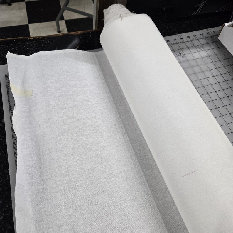 May include: A roll of white fabric, partially unrolled, lies on a cutting mat. The fabric appears to be a lightweight material, possibly cotton or linen. The scene suggests a sewing or crafting project.