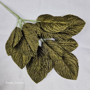 May include: A decorative stem featuring multiple dark olive green leaves with a textured, velvet-like finish. The leaves are attached to a thin, light green central branch. The text Timely Tresses is printed in the bottom left corner of the image.