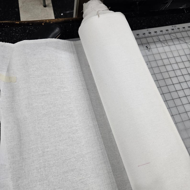 May include: A roll of white fabric sits next to a flat piece of the same material. The fabric appears to be a light, textured weave. A cutting mat with grid lines is visible in the background.