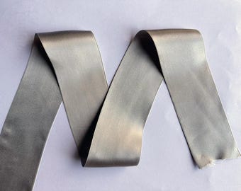 May include: A length of silver-gray satin ribbon, arranged in a wavy pattern. The ribbon has a smooth, reflective surface and a slightly frayed edge. Ideal for crafts, gift wrapping, or decorative purposes.