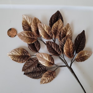 May include: A decorative branch featuring multiple fabric leaves in varying shades of metallic gold, bronze, and dark brown. The leaves have a textured, veined surface and are attached to a central stem. A copper penny is placed nearby for scale.