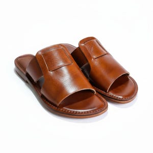 Può includere: Un paio di sandali in pelle marrone. I sandali hanno un design semplice con un cinturino largo e un piccolo dettaglio quadrato. Le suole sono marrone scuro. I sandali sono presentati su sfondo bianco. I sandali sono in pelle.