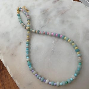 May include: A beaded necklace featuring a variety of pastel-colored beads. The necklace includes beads in shades of blue, pink, yellow, and gray. The clasp and chain are gold-colored. The necklace is displayed on a white marble surface.