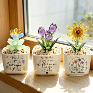 May include: Three white square ceramic pots containing glass flowers and succulents. The left pot says "Thank you for helping me grow Angela" with flowers. The middle pot says "Never forget the difference that you make". The right pot says "Teach them, love them, watch them grow Mrs. Katrina" with wildflowers.