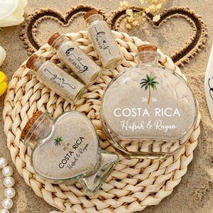 May include: A collection of clear glass bottles filled with sand, including a large round bottle and a heart-shaped bottle labeled COSTA RICA, Hafsah & Rayan, 20 26, and three smaller cylindrical vials with dates and initials printed on them.