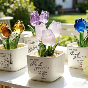 May include: Glass tulip flower arrangements in white square pots. The tulips are in shades of yellow, purple, pink, and blue. Each pot has a message of gratitude and a name. The pots are on a white surface.