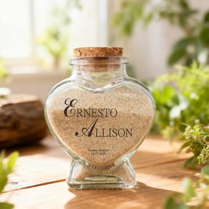 May include: A heart-shaped glass jar with a cork stopper, filled with light tan sand. The front of the jar features black text that reads Ernesto, Allison, Tucson Arizona, and the date 10.17.2025 printed directly onto the clear glass surface.