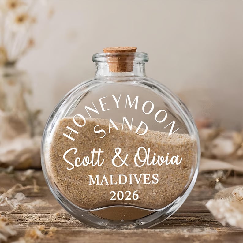 May include: A clear, round glass bottle with a cork stopper, partially filled with light tan sand. The front of the bottle features white text that reads HONEYMOON SAND, Scott & Olivia, MALDIVES, and 2026, printed in a mix of serif and script fonts.