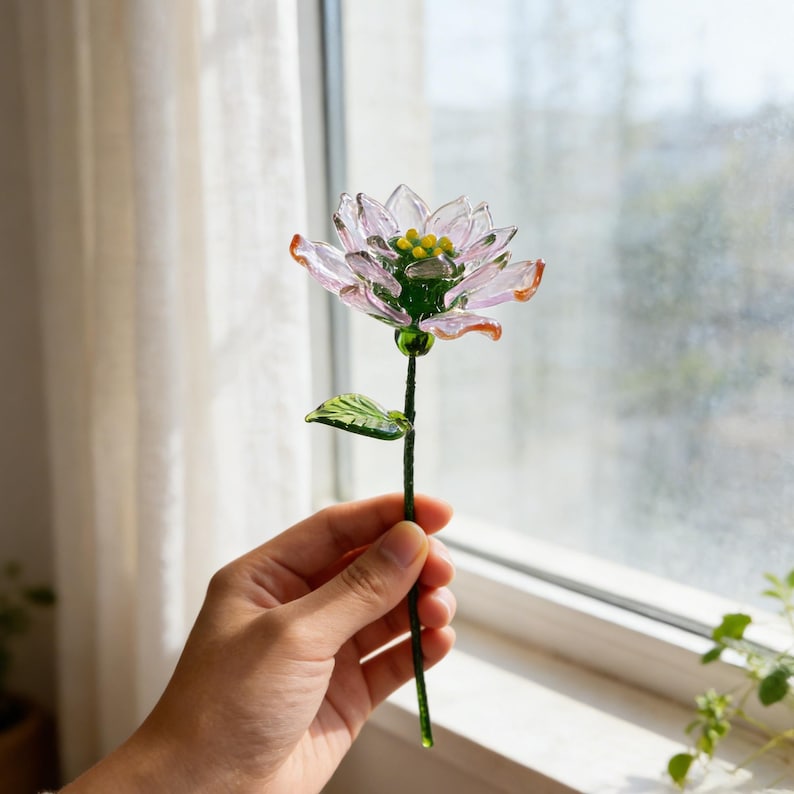 May include: A delicate glass flower with a pink and orange-tipped blossom, yellow center, and green stem and leaf. The handcrafted floral art piece is held in front of a window, showcasing its intricate details and translucent petals.