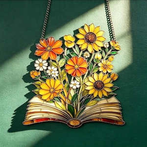Op de afbeelding: Een glas-in-lood zonnevanger met een open boek waaruit een boeket gele en oranje zonnebloemen en madeliefjes groeit. Het object hangt aan een metalen ketting en toont levendige bloemkleuren met donkere loden lijnen tussen de glasstukken.