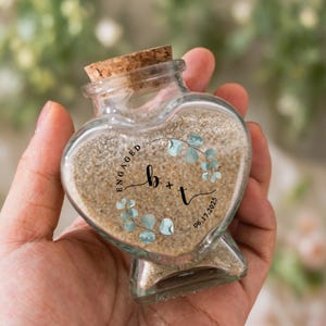 May include: A heart-shaped glass bottle with a cork stopper, filled with light tan sand. The front features a black printed design with the letters b + t, the word ENGAGED, the date 06.17.2025, and a decorative wreath of light blue eucalyptus leaves.