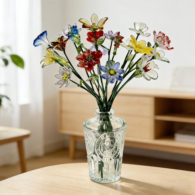 May include: A clear glass vase with a detailed pattern holds a bouquet of colorful glass flowers. The flowers feature red, yellow, blue, and white petals with green stems. The vase sits on a light-colored wooden table.