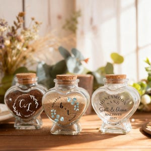 May include: Three heart-shaped glass jars with cork stoppers, each filled with sand. The jars feature custom engravings: one with 'C+L', one with 'b+t' and eucalyptus leaves, and one with 'Honeymoon Sand Scott & Olivia Maldives 2025'.