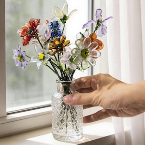 May include: A small glass vase filled with colorful, artificial flowers. The flowers are various colors, including red, white, blue, and orange. The vase has a diamond pattern and is held by a hand near a window.