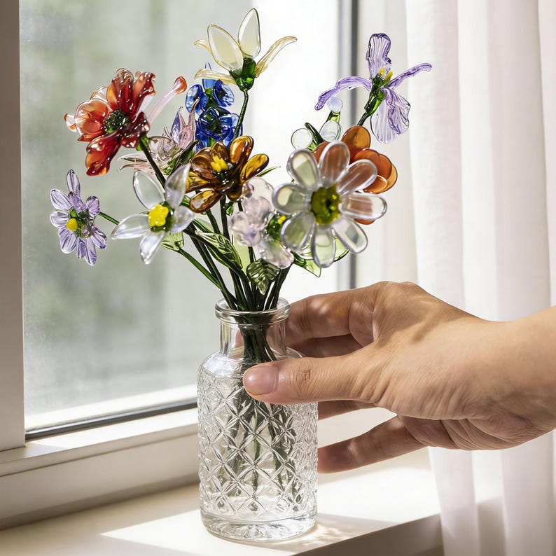 May include: A small glass vase filled with colorful, artificial flowers. The flowers are various colors, including red, white, blue, and orange. The vase has a diamond pattern and is held by a hand near a window.