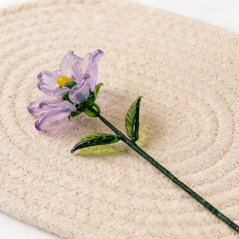 May include: A handcrafted glass flower with a purple blossom and a yellow center. The flower has green leaves and a green stem. The flower is resting on a beige woven mat.