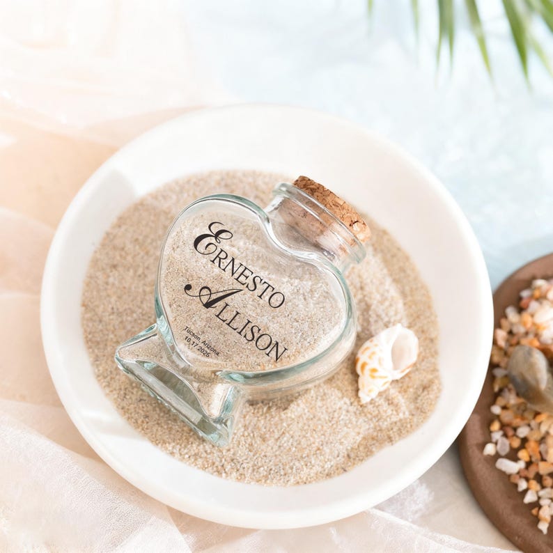 May include: A heart-shaped glass bottle with a cork stopper, filled with sand and a small seashell. The glass is engraved with the names Ernesto and Allison, along with the location Tucson, Arizona and the date 10.17.2026 in black lettering.