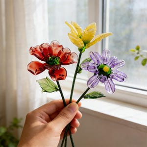 May include: A bouquet of three glass flowers in red, yellow, and purple. The flowers have green stems and leaves. The red flower is a poppy, the yellow flower has six petals, and the purple flower has multiple petals.