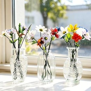 May include: Three clear glass vases with colorful artificial flowers sit on a white windowsill. The flowers include daisies, roses, and other blooms in shades of red, yellow, white, and purple. The vases have different etched designs.