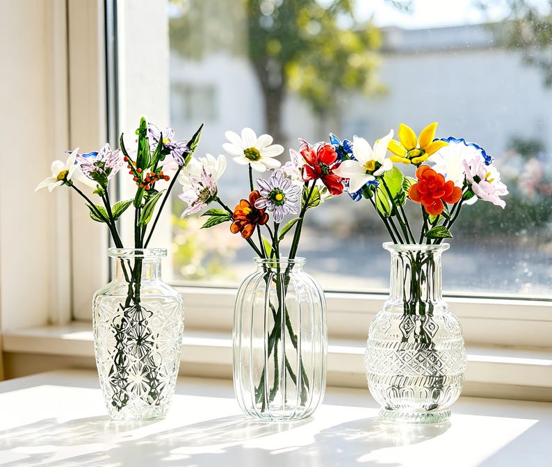May include: Three clear glass vases with colorful artificial flowers sit on a white windowsill. The flowers include daisies, roses, and other blooms in shades of red, yellow, white, and purple. The vases have different etched designs.