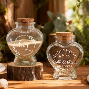 May include: Two heart-shaped glass jars with cork stoppers. The left jar contains sand and is engraved with "CARLSON & LINDSAY", "C+L", and "AUGUST 29, 2025". The right jar is engraved with "HONEYMOON SAND", "Scott & Olivia", "MALDIVES", and "2025".