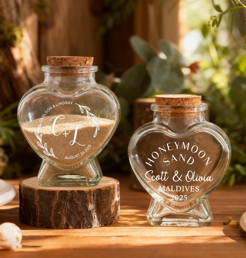 May include: Two heart-shaped glass jars with cork stoppers. The left jar contains sand and is engraved with "CARLSON & LINDSAY", "C+L", and "AUGUST 29, 2025". The right jar is engraved with "HONEYMOON SAND", "Scott & Olivia", "MALDIVES", and "2025".