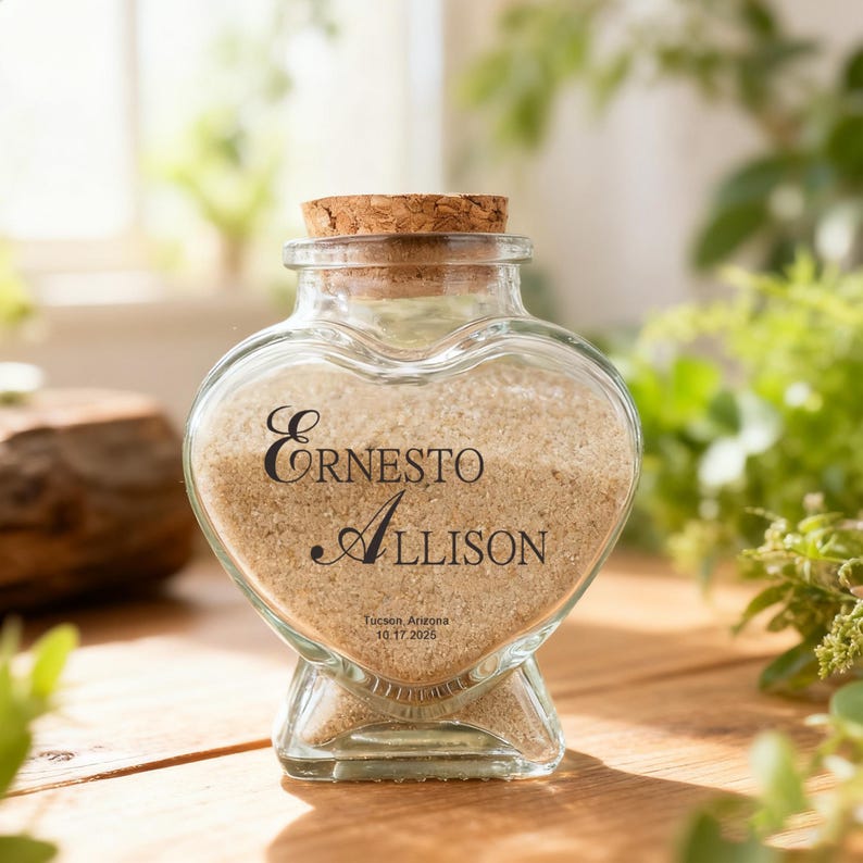 May include: A heart-shaped glass jar with a cork stopper, filled with light tan sand. The front of the jar features black text that reads Ernesto, Allison, Tucson Arizona, and the date 10.17.2025 printed directly onto the clear glass surface.