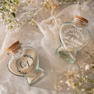 May include: Two heart-shaped glass bottles filled with sand and cork stoppers. One bottle features white text reading HONEYMOON SAND, Scott & Olivia, MALDIVES 2025. The other features black text C+L, CARLSON & LINDSAY, AUGUST 29 2025 with leaf accents.