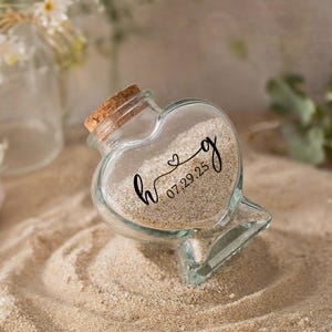 May include: A clear glass heart-shaped bottle with a cork stopper, filled with light beige sand. The front of the glass features black printed text that reads h, a small heart symbol, g, and the date 07.29.25 in a cursive script font.