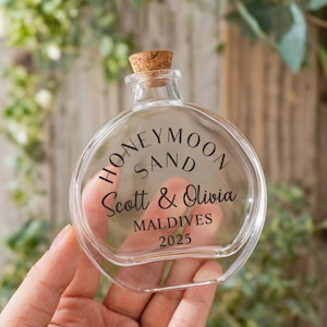 May include: A clear, round glass bottle with a cork stopper. The front of the bottle features black text that reads HONEYMOON SAND, Scott & Olivia, MALDIVES, and 2025. The bottle has a short neck and a flat, circular body design for display.
