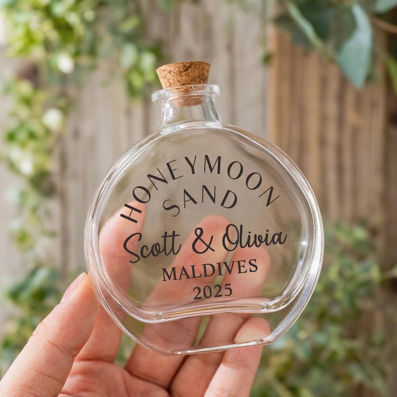 May include: A clear, round glass bottle with a cork stopper. The front of the bottle features black text that reads HONEYMOON SAND, Scott & Olivia, MALDIVES, and 2025. The bottle has a short neck and a flat, circular body design for display.
