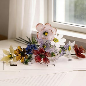 May include: A colorful glass flower bouquet featuring various flower types in shades of pink, yellow, blue, red, and white. The stems are dark green. The flowers are arranged on a white surface.