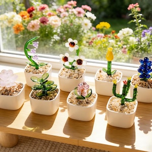 May include: A selection of glass flower sculptures in small white square pots. The flowers include a lily of the valley, daisies, a tulip, a hyacinth, a cactus, a succulent, and a rose. The sculptures are on a wooden table near a window.