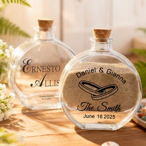 May include: Two round clear glass bottles with cork stoppers. The left bottle features the text Ernesto Allis, Tucson, Arizona, 10.17.2025. The right bottle contains sand and features the text Daniel & Gianna, The Smith, June 16 2025, and two rings.